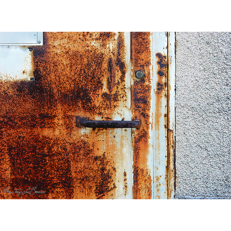 Ancienne porte métallique très rouillée avec poignée sombre, peinture claire résiduelle et larges plages de corrosion orange, brune et noire, œuvre photographique de Pierre Tomy Le Boucher.