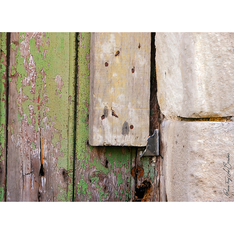 Détail d’une ancienne porte en bois à la peinture verte très écaillée, avec fissures, anciennes fixations et réparation en bois clair contre un mur de pierre, œuvre photographique de Pierre Tomy Le Boucher.