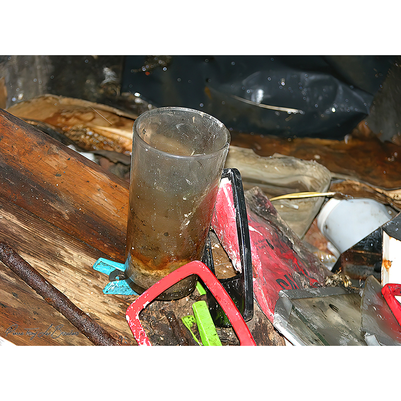 Verre cylindrique abandonné, rayé, terni et couvert de dépôts, entouré de bois, de métal et de fragments colorés, œuvre photographique de Pierre Tomy Le Boucher.