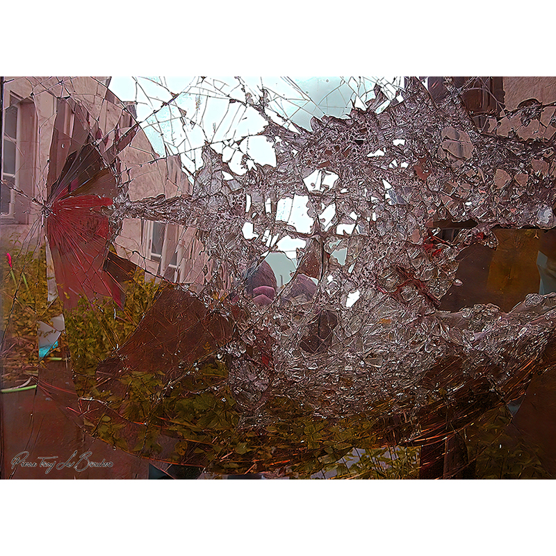 Surface de vitre brisée formant un réseau dense de fissures et d’éclats, avec des reflets et des transparences déformées laissant apparaître l’environnement derrière, œuvre photographique de Pierre Tomy Le Boucher.