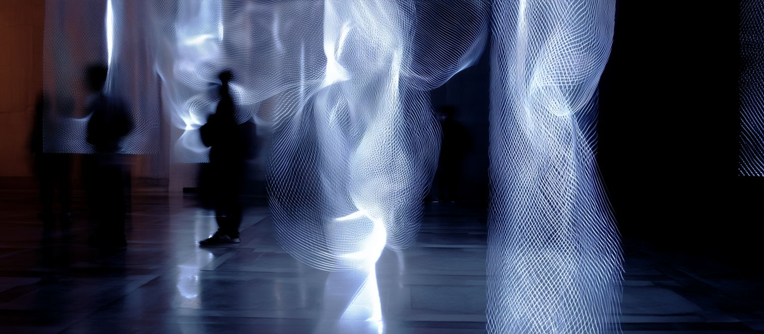 Silhouette humaine devant une installation lumineuse abstraite composée de flux de lumière ondulants évoquant le temps réel et le mouvement.